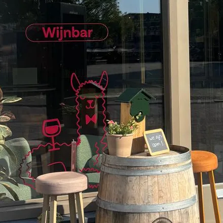 De lama met een glas wijn op het raam van de wijnbar, met een wijnton en krukken ervoor
